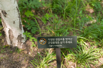 A sign in a foreign language warns people not to walk on the grass. The sign is placed in a field with trees and grass