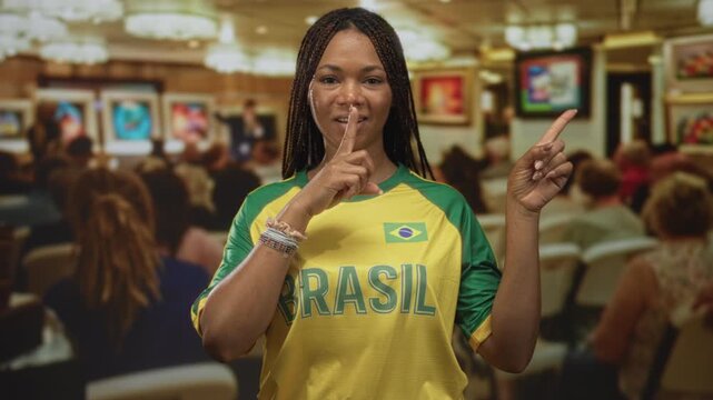 Woman with finger to lips for silence pointing at artwork in a building gallery wearing yellow brasil jersey and bracelets; quiet reflection.
