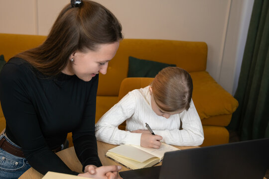 Mother and daughter are studying together at home, with the mother helping the daughter with her homework while the child writes in a notebook, creating a supportive learning environment