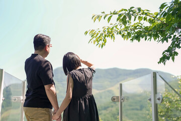 A couple is standing on a balcony overlooking a beautiful mountain range. They are holding hands and looking out at the view. Scene is peaceful and romantic