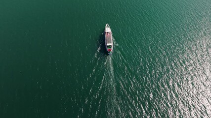 Top-down drone descends toward a moving ferry, showing its wake cutting across calm green water under bright daylight.