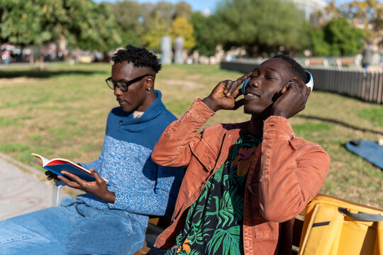 Two young African American men relaxing in a park, one listening to music, the other reading a book