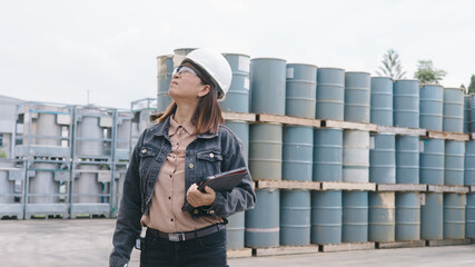 A woman wearing a hard hat and glasses observes a storage area filled with large barrels. She holds a clipboard and appears focused on her work in an industrial environment