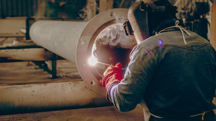 A worker is welding a large metal pipe inside a workshop in the evening. Sparks and smoke are visible as the welder focuses on the task at hand, wearing protective gear