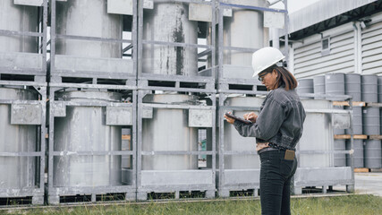 A worker wearing a hard hat and safety glasses checks a tablet while standing in front of large metal storage tanks at an industrial facility during daylight