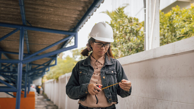 A woman wearing a hard hat and safety glasses carefully measures a wall with a tape measure. She stands in a construction area surrounded by greenery and buildings, focusing on her task