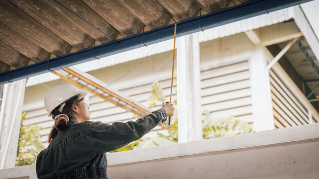 A construction worker wearing a hard hat and safety glasses measures a space with a tape measure inside a building under construction