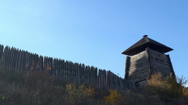 Ancient wooden fortress palisade with watchtower on a hill under evening sky.