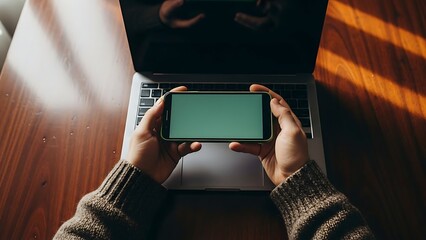 Hands holding a smartphone with a green screen above a laptop on a wooden table.
