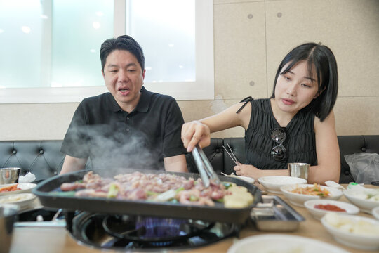 A man and a woman are sitting at a table with a hot plate of food in front of them. The man is cooking the food, while the woman looks on. Scene is one of warmth and togetherness - Powered by Adobe