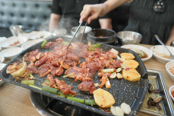 A person is cooking meat on a grill. The meat is being cooked with vegetables and there is a lot of steam coming from the grill