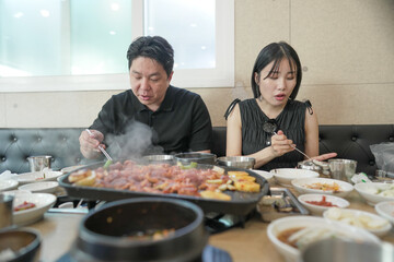 A man and woman are eating at a restaurant. The man is eating a dish with a lot of meat and vegetables. The table is full of food and there are many bowls
