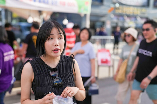 A woman in a black tank top is holding a bag and looking at something. She is surrounded by other people, some of whom are carrying handbags. The scene appears to be a busy street or market