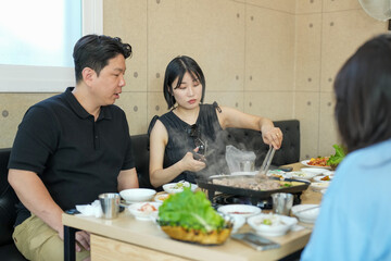 A man and a woman are sitting at a table with a lot of food in front of them. The woman is cooking something on a hot plate. Scene is casual and relaxed, as the people are enjoying a meal together