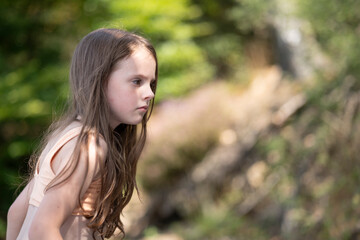 Naklejka premium Side view of thoughtful girl waist up with blurred green foliage background, emotional childhood outdoor portrait with copy space
