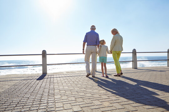 Grandparents, grandchild and holding hands on beach promenade for walk, ocean view and bonding together. Back, grandma and grandpa with kid outdoor by sea for sightseeing, family connection or space.