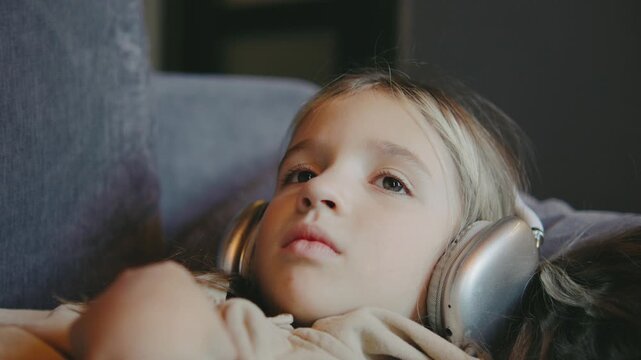 Sad little girl with headphones listening to music on a couch, lost in her thoughts in a cozy living room during a quiet afternoon