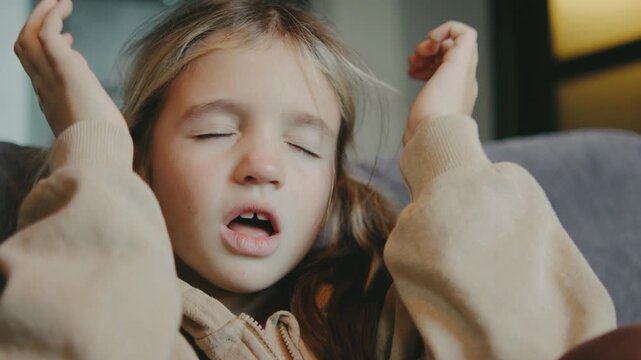A young girl sits on a couch with a distressed expression, covering her face with her hands. She seems overwhelmed and sad in a soft-lit room.