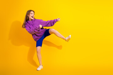 Young woman in purple hoodie kicks with joy against vibrant yellow background