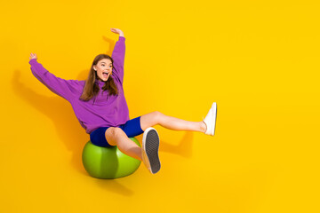 Young woman in purple hoodie and blue shorts sits on a green exercise ball leaps with joy against a bright yellow background for a playful fitness lifestyle image