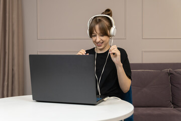 Young woman wearing headphones is using laptop at home, studying online and expressing joy and excitement for successful e-learning experience