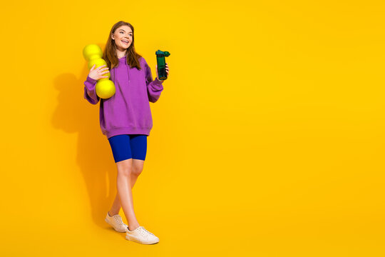 Young woman in a purple hoodie and blue shorts poses with dumbbells and a bottle against a bright yellow background - Powered by Adobe