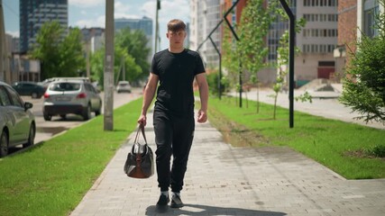 youthful man strolling, white young man with gym bag walking urban street, confident young man in casual attire exploring vibrant summer urban environment with parked vehicles and green spaces