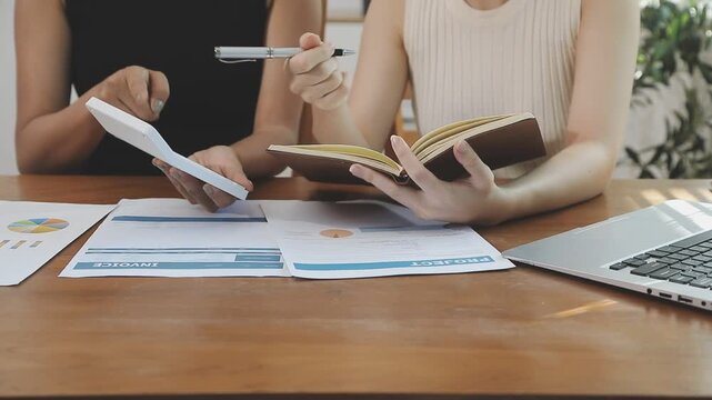 Two financial accountants analysing business spreadsheets on laptop computer, sitting at office desk, looking through paper documents, using calculator working with databases, checking excel tables