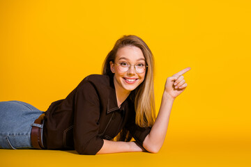 Young female student lying on yellow background smiling and pointing to the side