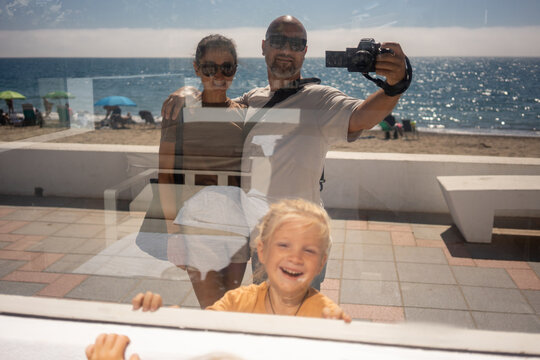 Reflection of smiling family with camera in glass window by beach on sunny summer day