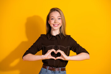 Young woman in casual fashion smiles and forms a heart with hands against bright yellow background...
