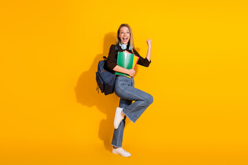Young woman with backpack and book jumps with joy in a bright yellow studio background