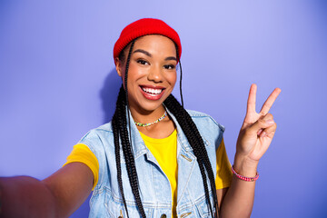 Young woman in red beanie denim vest smiling and flashing peace sign against violet background for vibrant fashion lifestyle stock image