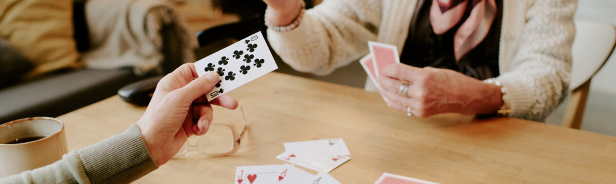 Senior Caucasian woman and senior Caucasian man playing card game at table, hands holding playing cards, coffee mug on wooden surface, engaging in leisure activity together