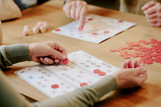 Senior Caucasian woman and senior Caucasian man playing bingo at table, hands placing red chips on bingo cards, engaging in leisure activity, close up of hands and game pieces