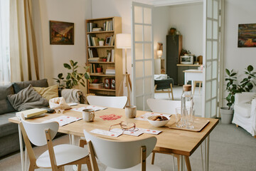 Modern dining room featuring wooden table set with snacks, drinks, and playing cards, cozy living area with sofa and bookshelves in background, bright and inviting space