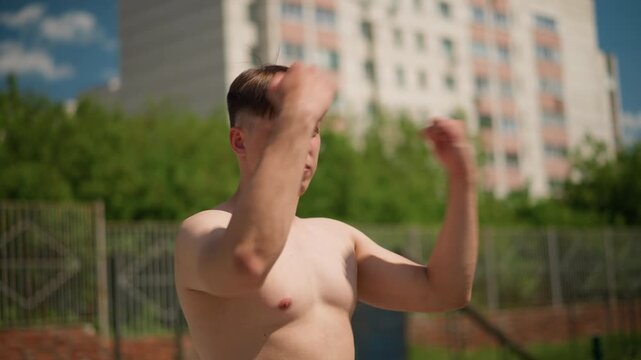 unclothed man works on punches, white male exercises punch techniques near city greenery backdrop, man without shirt performs punching drills in front of apartment buildings and lush trees