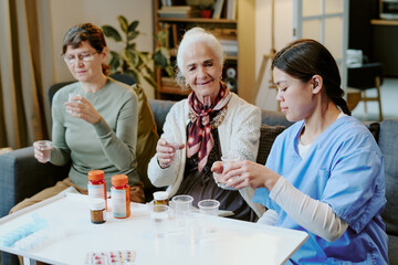Senior Caucasian woman sitting between middle aged Caucasian woman and young adult Hispanic woman in scrubs, all holding cups and preparing to take medication at home