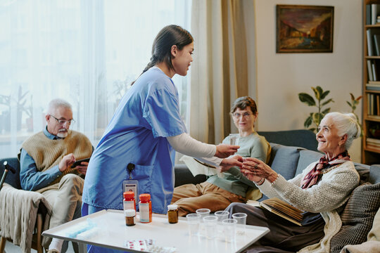 Middle aged Hispanic female nurse handing medication to senior Caucasian woman sitting on sofa, while middle aged Caucasian man and woman sitting nearby engaging in conversation