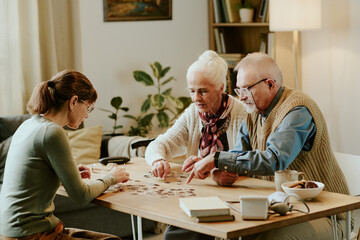 Obraz premium Senior Caucasian woman and senior Caucasian man sitting at table assembling puzzle with middle aged Caucasian woman, all focused on activity together in cozy home setting