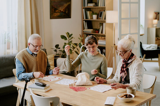 Three seniors, two Caucasian women and one Caucasian man, sitting at table playing bingo together, holding bingo chips and cards, engaging in group leisure activity in living room