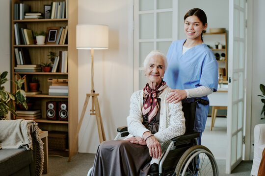 Senior Caucasian woman sitting in wheelchair with disability, smiling while young adult Hispanic female nurse standing behind her, both looking at camera in home setting - Powered by Adobe