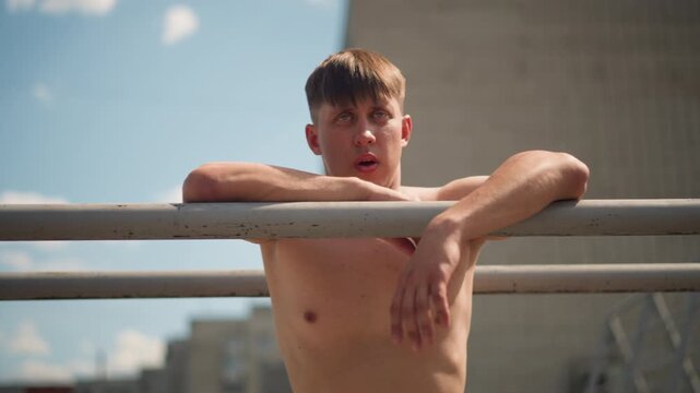 relaxed man by city railing, barechested individual leans against railing with city view behind, naked male subject leans nonchalantly on metal barrier with cityscape reflected in his face