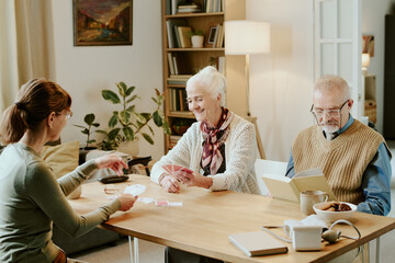 Obraz premium Senior Caucasian woman smiling while holding playing cards, sitting at table with senior Caucasian man reading book and young adult Caucasian woman dealing cards indoors