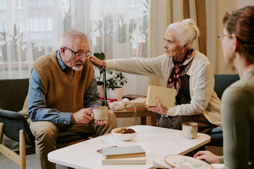 Senior Caucasian woman comforting senior Caucasian man while holding book, sitting together in living room with another middle aged Caucasian woman listening nearby