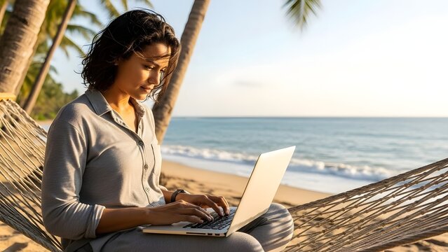Young woman with dark curly hair working on a laptop computer while relaxing in a hammock on a sunny tropical beach near the ocean