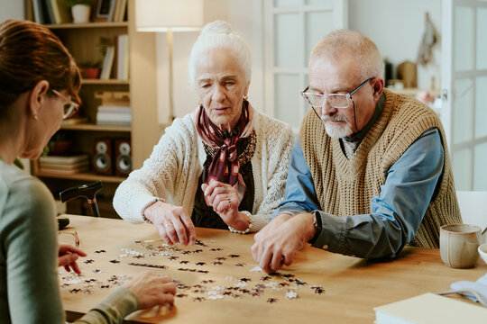Senior Caucasian woman and senior Caucasian man sitting at table assembling jigsaw puzzle with middle aged Caucasian woman, all focusing on puzzle pieces and collaborating together