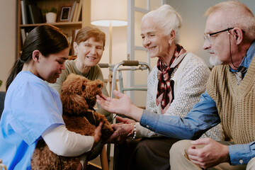 Senior Caucasian woman smiling while petting dog held by young adult Hispanic female caregiver, middle aged Caucasian woman and senior Caucasian man sitting nearby interacting with animal