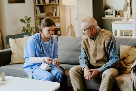 Hispanic young adult female nurse demonstrating blood pressure monitor to Caucasian senior man sitting on sofa in home setting, both focusing on medical device during healthcare visit