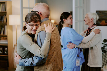 Senior Caucasian woman hugging senior Caucasian man while smiling, young Hispanic female nurse supporting senior Caucasian woman in background, showing caregiving interaction
